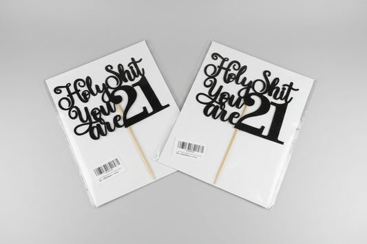 Two 'Holy Shit You Are 21' cake toppers on packaging with a neutral background.